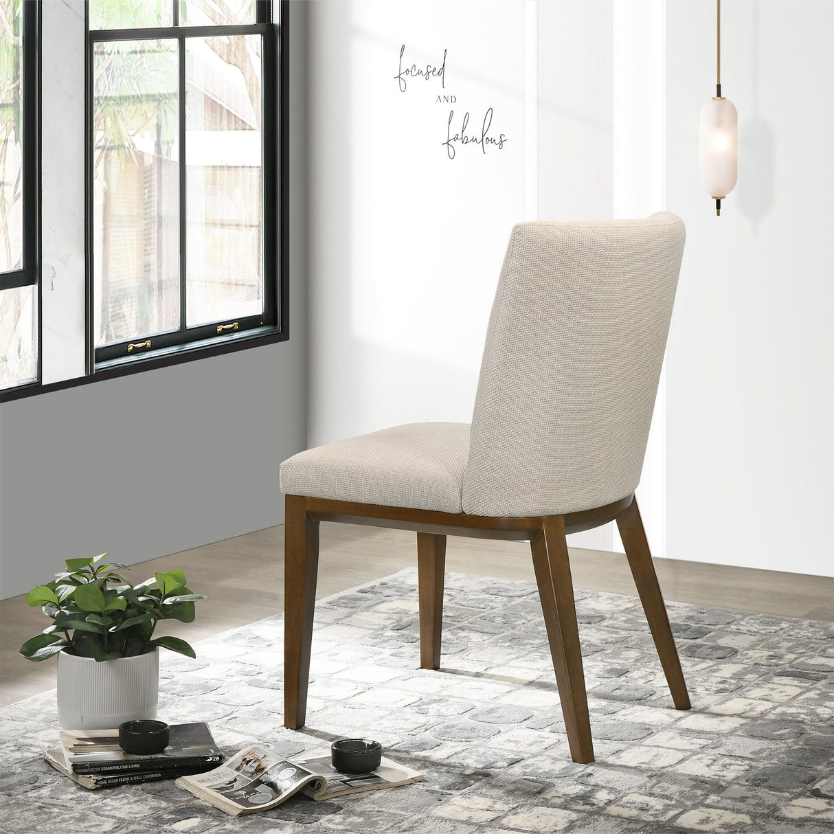 An Elmwood Stylish Dining Chair in beige with wooden legs is on a patterned rug. A potted plant, magazines, and an earbud case are placed nearby. A modern pendant light illuminates the room where "Focused and Fabulous" adorns the wall.