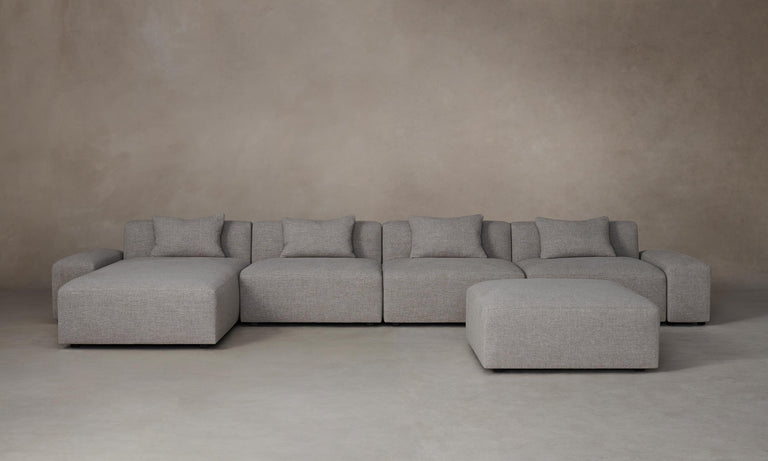 The Breuer Modular Chaise Armless Sectional 114" in Belgian Linen Agate, a spacious and gray-toned modular sectional sofa with multiple cushions, is arranged on a neutral-toned floor against a plain beige wall. The setup includes a matching ottoman positioned slightly to the right.