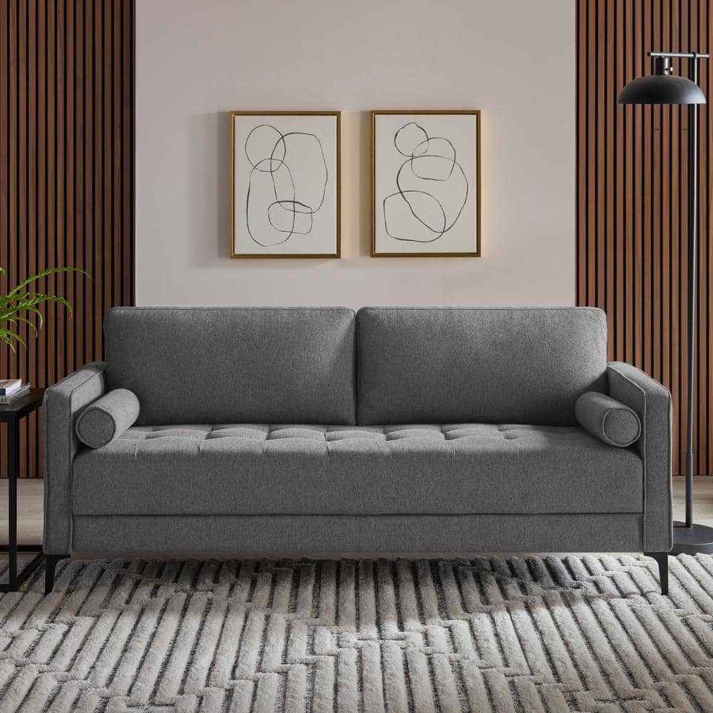 A modern living room featuring the StyleWell Goodwin Mid-Century Modern Sofa with throw pillows on a patterned rug. The wall behind showcases two abstract line drawings and is accented with vertical wooden panels. A tall black floor lamp and a small side table with a plant complete the scene.
