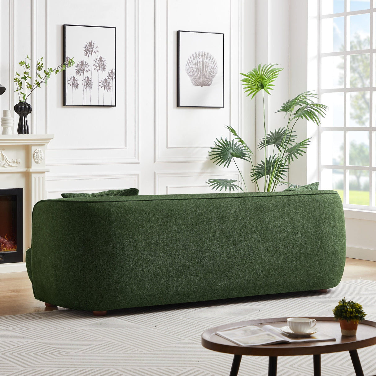 Facing large windows, a modern living room features a Larisa Mid-Century Modern Linen Sofa. Minimalist decor includes framed botanical prints, a potted plant, and a round wooden coffee table with a book and teacup, all harmonized by neutral walls and flooring.
