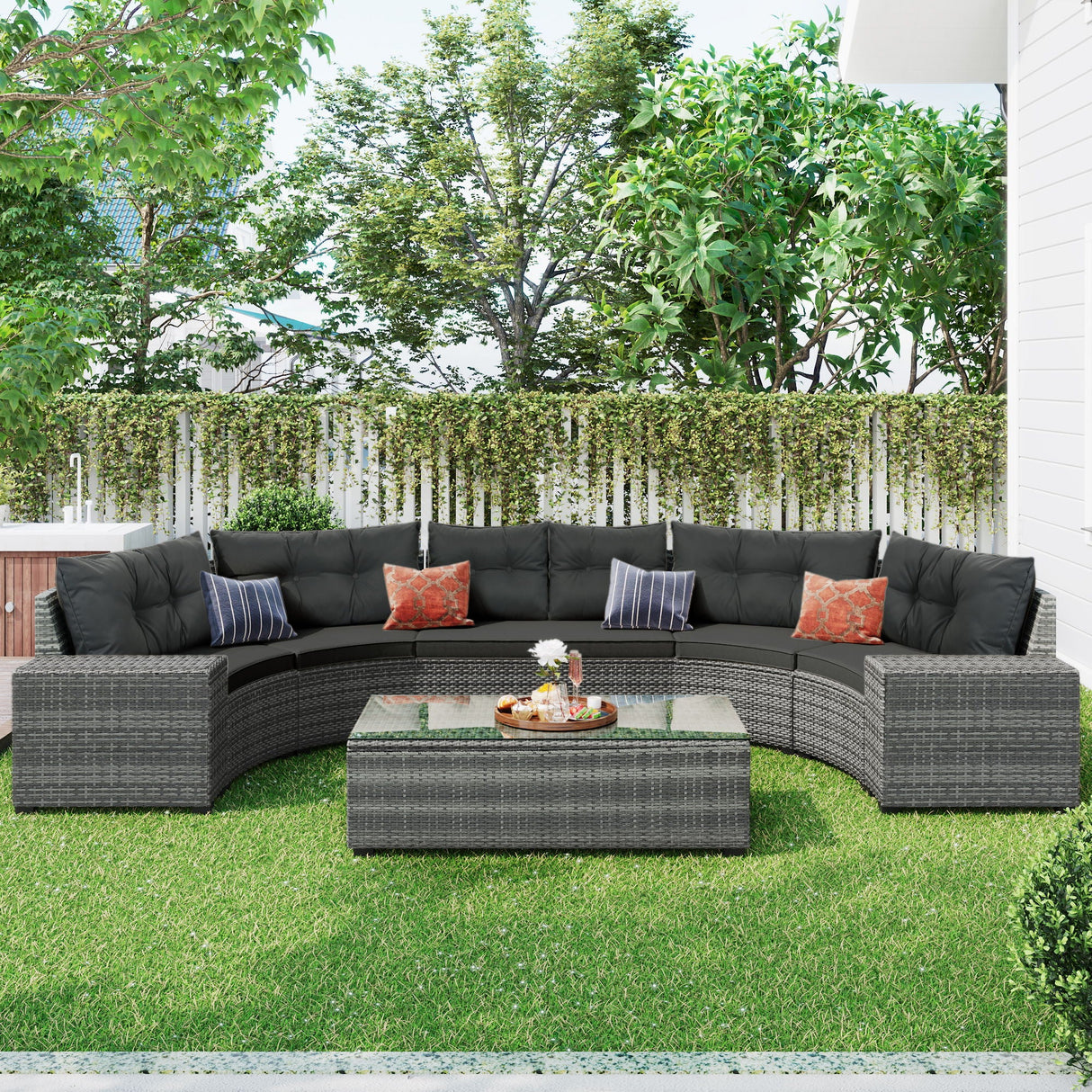 The outdoor patio features an 8-piece wicker round sofa set with a curved gray sectional and black cushions, adorned with orange-patterned pillows. A matching rectangular wicker coffee table holds a tray with books and a small plant, complementing the green lawn, white fence, and trees for added coziness.