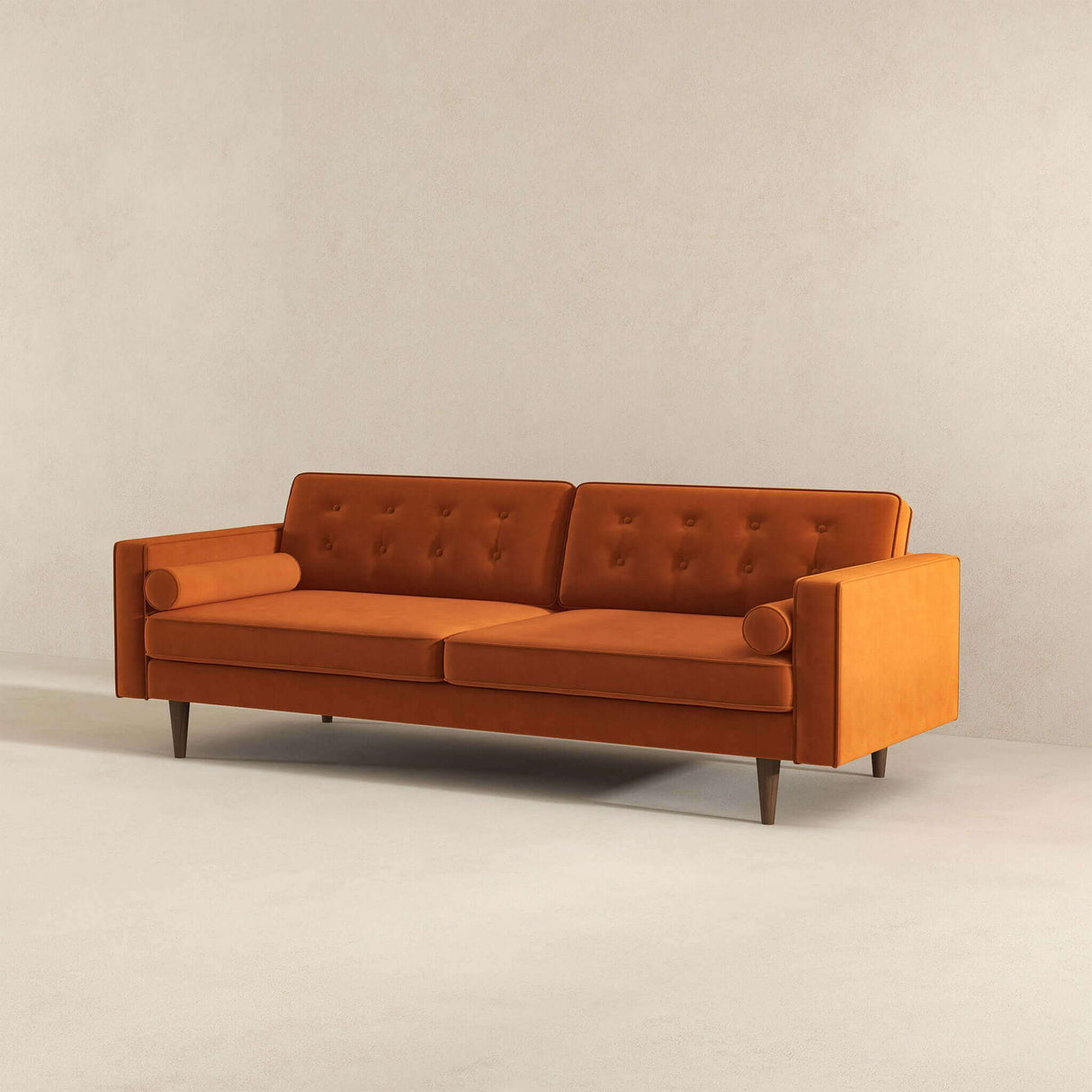 The Casey Mid Century Modern Sofa features a mid-century design with an orange finish, two cushions, bolster pillows, a button-tufted backrest, and wooden legs. It is set against a plain beige wall and floor.
