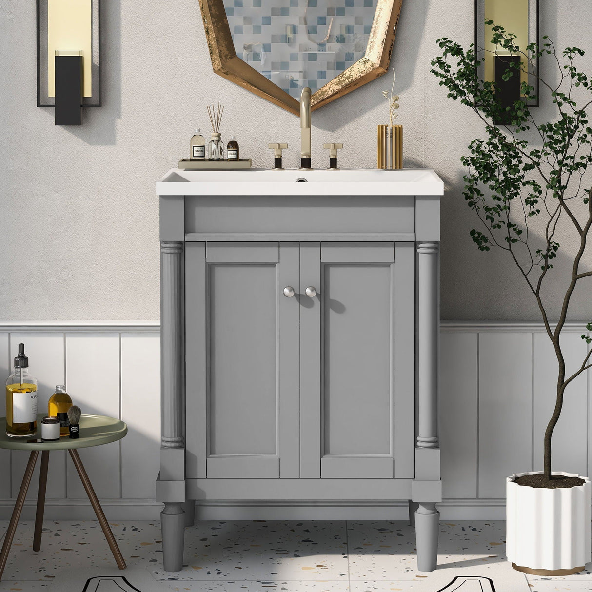 The Bathroom Vanity With Top Sink features a grey cabinet and white sink with a faucet. An octagonal mirror in gold trim hangs above, flanked by wall sconces. A table with toiletries sits to the left and a potted plant to the right.