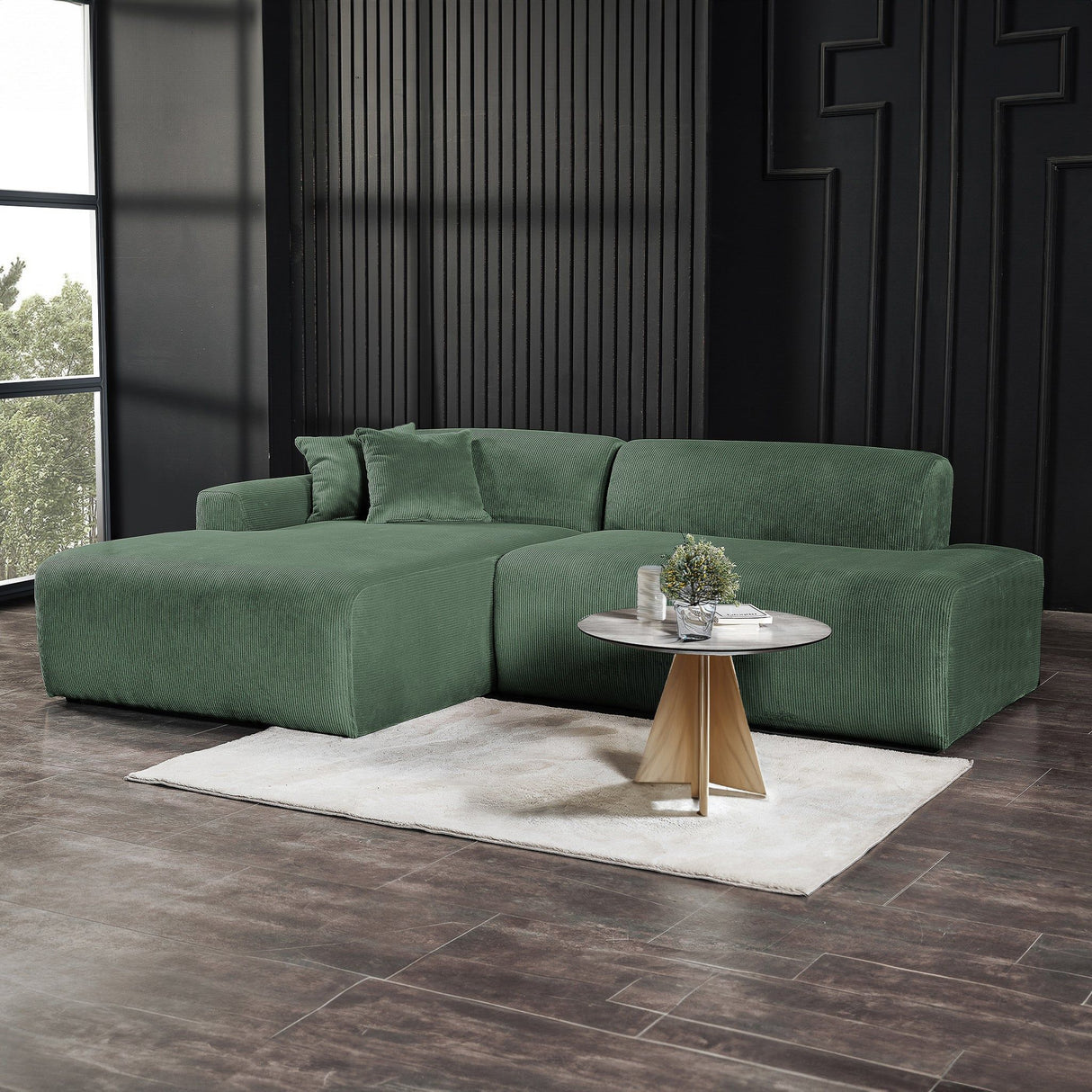 A modern living room featuring the Mar High-Quality Sectional Sofa in deep green, paired with a small circular table holding a potted plant. The room boasts dark wood floors, grooved walls, a large window for natural light, and a white rug positioned under the table.