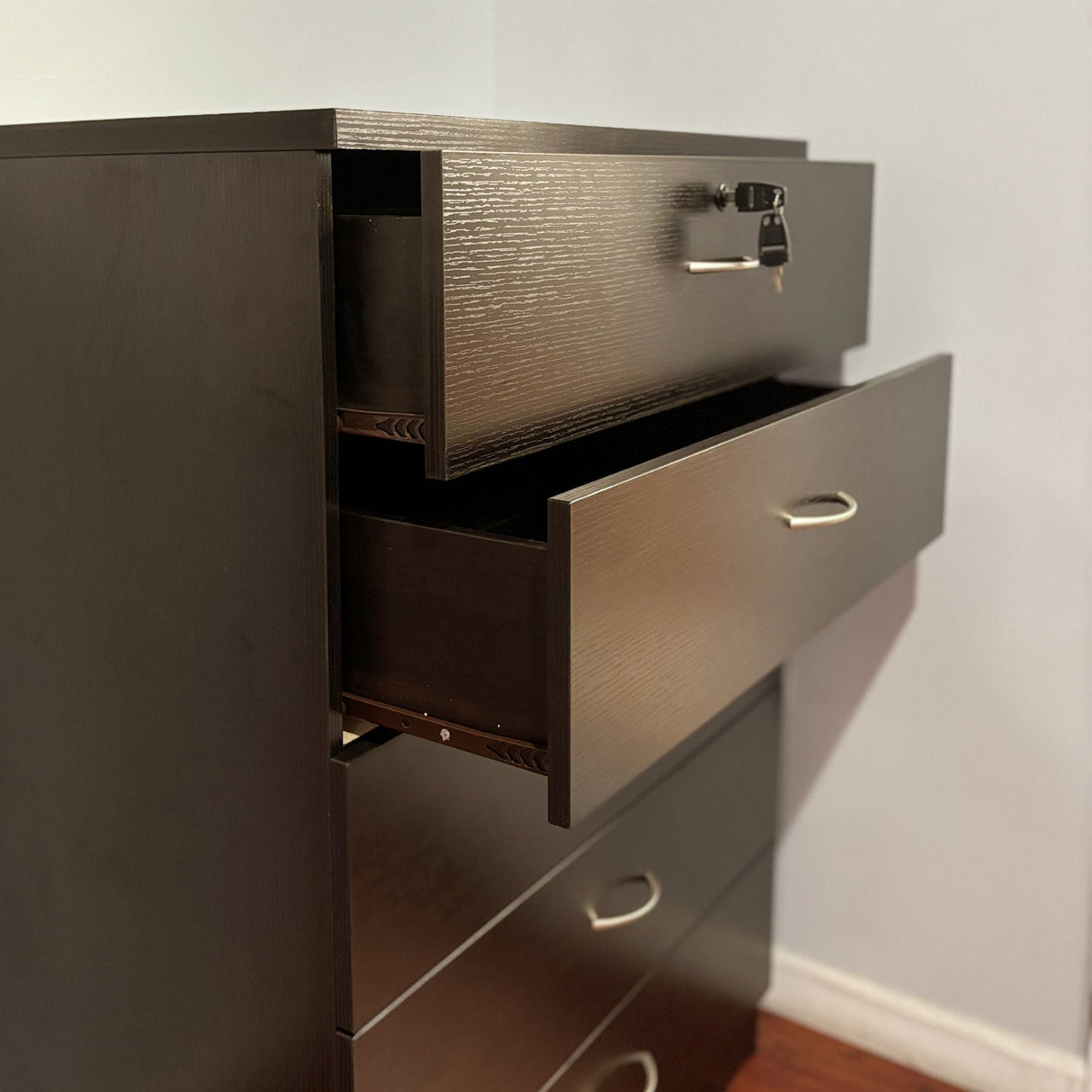 A 5 Drawer Chest with a melamine laminate and metal handles stands on a hardwood floor against a plain wall. The top drawer, equipped with metal glide rails and a locking mechanism, is slightly open with a key inserted, while the other three drawers remain closed.