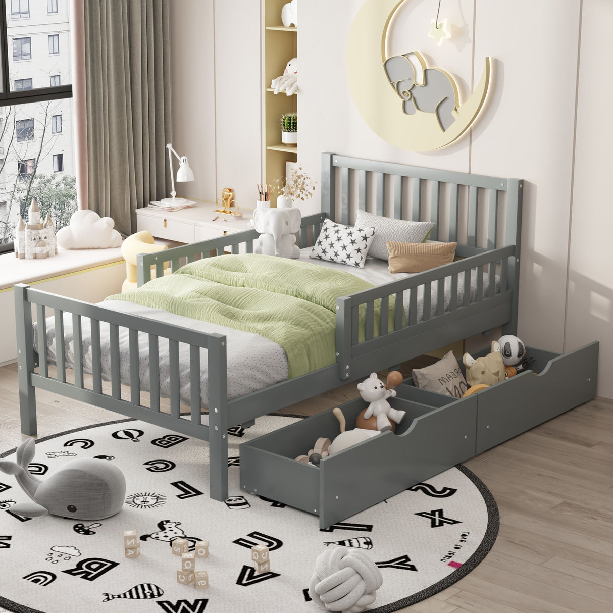 A child's room features a gray platform bed with guardrails, a green blanket, and stuffed toys. The bed's two storage drawers hold more toys. A moon-shaped light hangs above, while shelves with books and toys line the wall. Natural light flows through the window onto a round rug on the wooden floor.