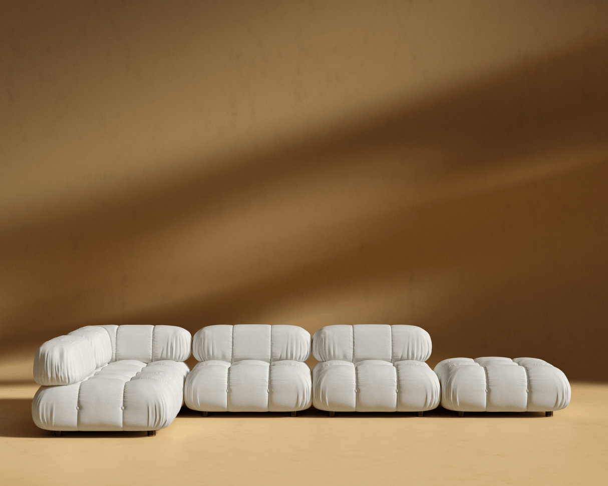 The Belia Modular Sectional in white is showcased against a light brown wall. It boasts a modern, tufted design with multiple pieces, including a chaise lounge, three seats, and an ottoman. The scene is brightly lit with soft shadows.