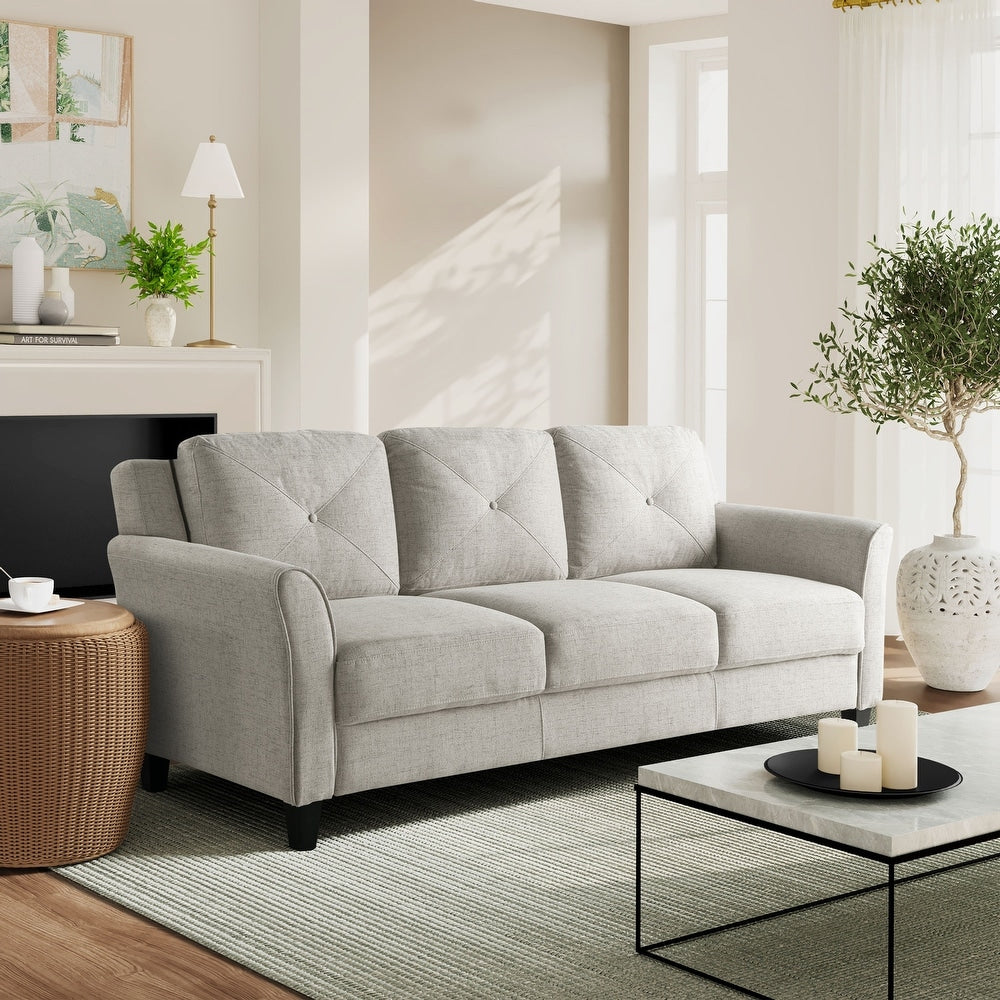 A modern living room with an iLounge Harvard Microfiber Sofa in light gray, a wicker side table, and a glass coffee table adorned with candles. The space is complemented by a potted plant, a table lamp, framed wall art, and large windows allowing ample natural light.