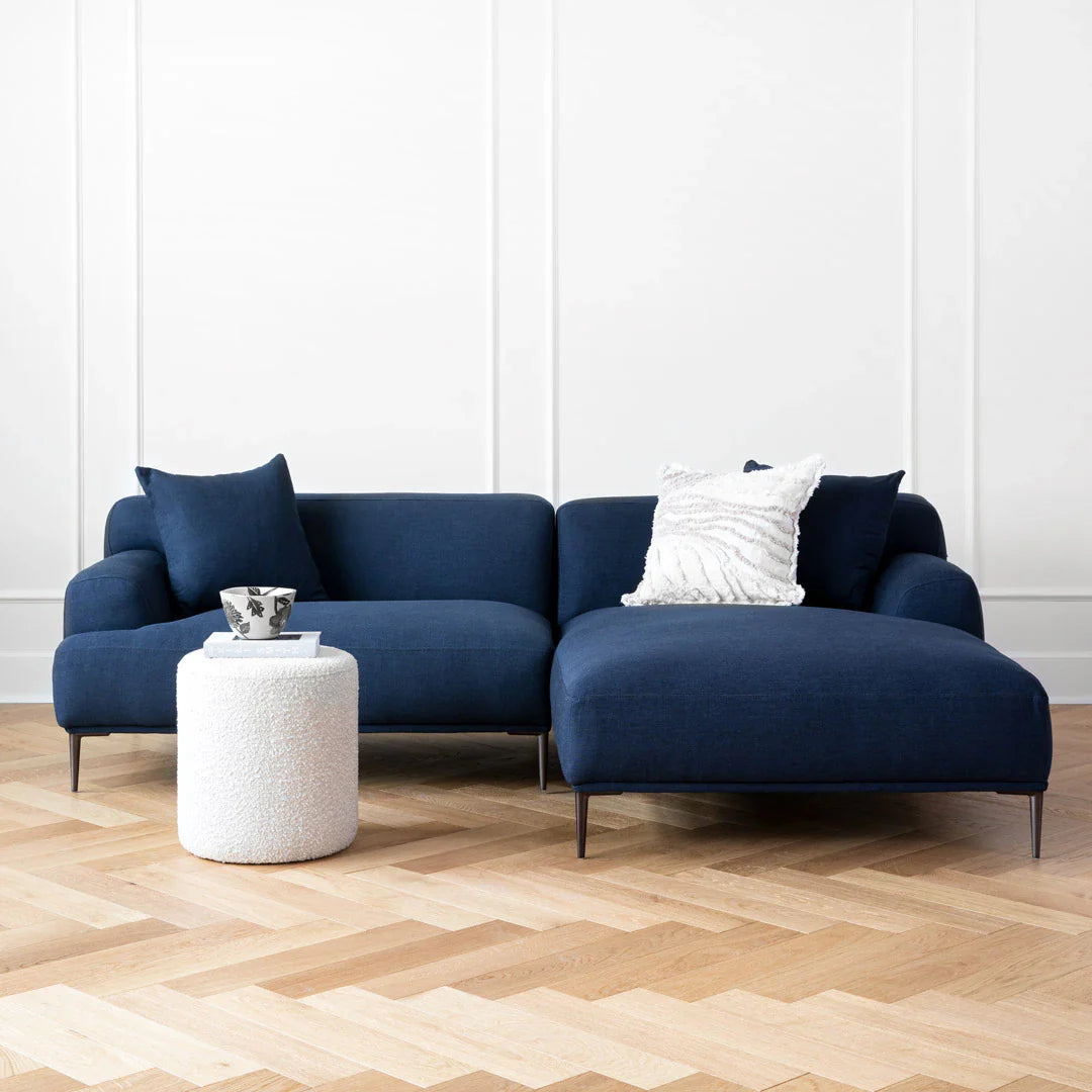 The living room is styled with the Abisko 94.5 Right Sectional in Aurora Blue, which includes two cushions. In front of the sectional, there's a small white textured cylindrical side table featuring a decorative bowl. The space is completed with hardwood flooring and plain white walls.