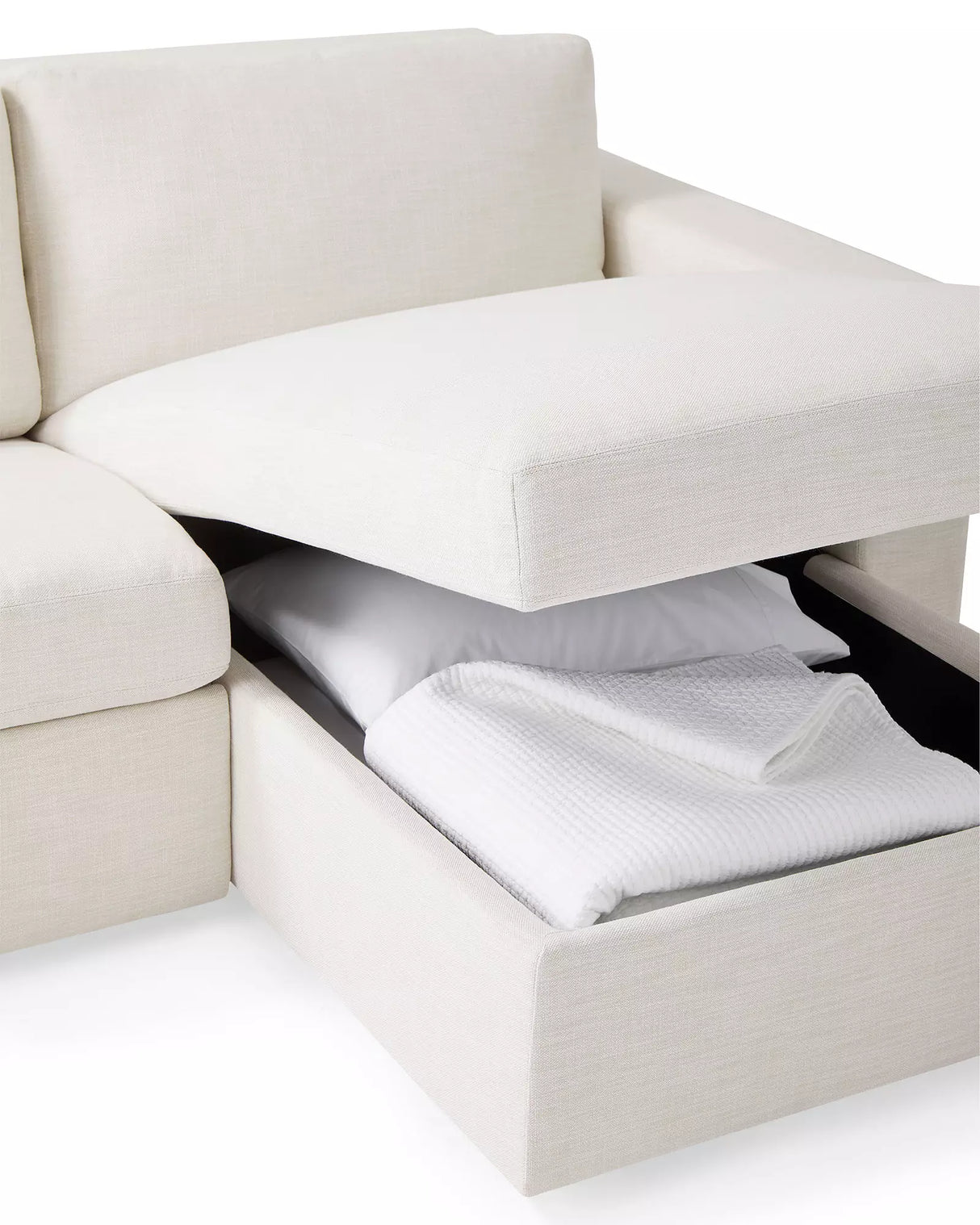 The Conover Sleeper Chaise Sectional in beige features a storage compartment beneath the seat cushion, revealing a folded white blanket and pillow that highlight its multifunctional design.