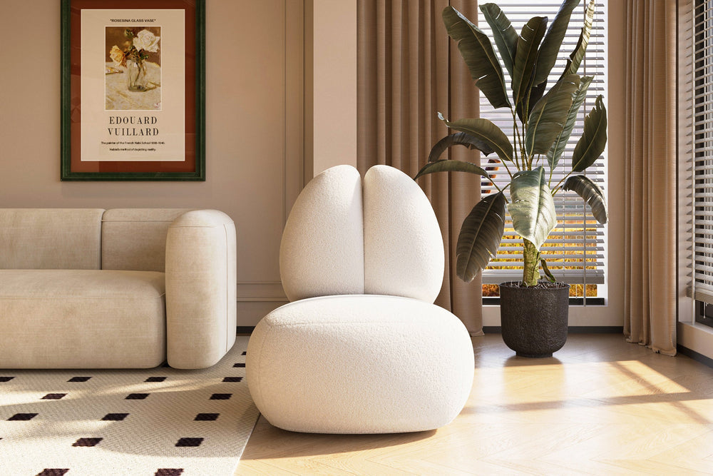A modern living room with the Moony Fleece Curved Lounge Chair by Acanva in white, paired with a beige sofa on a patterned rug. Beside the chair stands a large plant in a pot. A framed poster hangs on the beige wall, and natural light streams through the window with blinds.