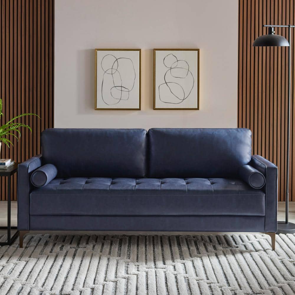 A modern living room showcases the StyleWell Goodwin Mid-Century Modern Sofa with its navy blue tone and cylindrical cushions, positioned against a light-colored wall with wooden panels. Above the sofa, two abstract line art frames are displayed, while a floor lamp stands on a patterned rug nearby.