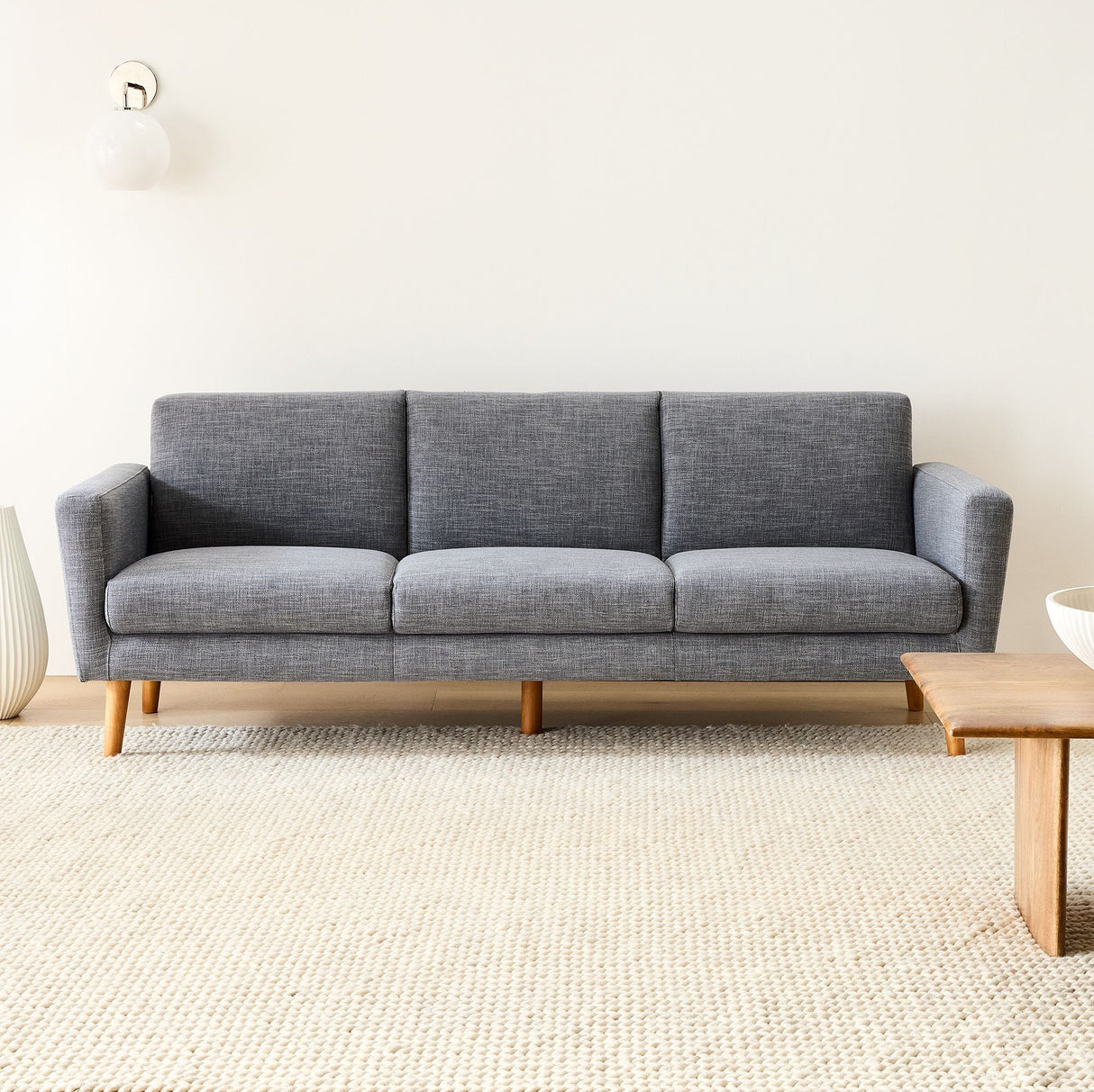A modern living room showcases the Oliver Sofa, a gray three-seater with wooden legs, placed on a cream woven rug. A wooden coffee table and a decorative vase are visible against a white wall with a simple light fixture.