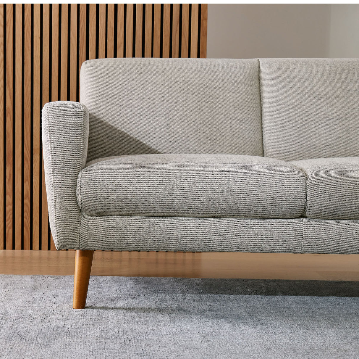 The Oliver Sofa, featuring light gray upholstery and wooden legs, is displayed in a living room. The backdrop includes a wooden slat wall, while a light gray carpet covers the floor, showcasing a minimalist, contemporary design style.