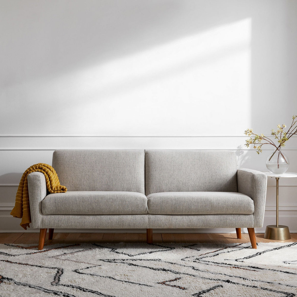 In a contemporary living room, the Oliver Sofa in light gray with wooden legs sits against a white wall. A mustard yellow blanket is elegantly draped over its armrest. Beside the sofa, there's a round side table adorned with a vase of branches. A patterned area rug decorates the floor.