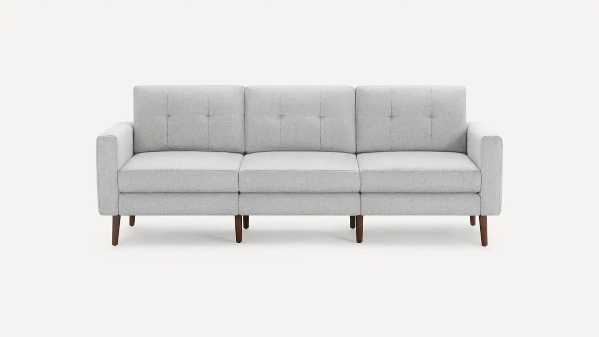 The Nomad Fabric Sofa is a contemporary gray piece with a minimalist design, featuring three cushions and elegant wooden legs, set against a simple backdrop.