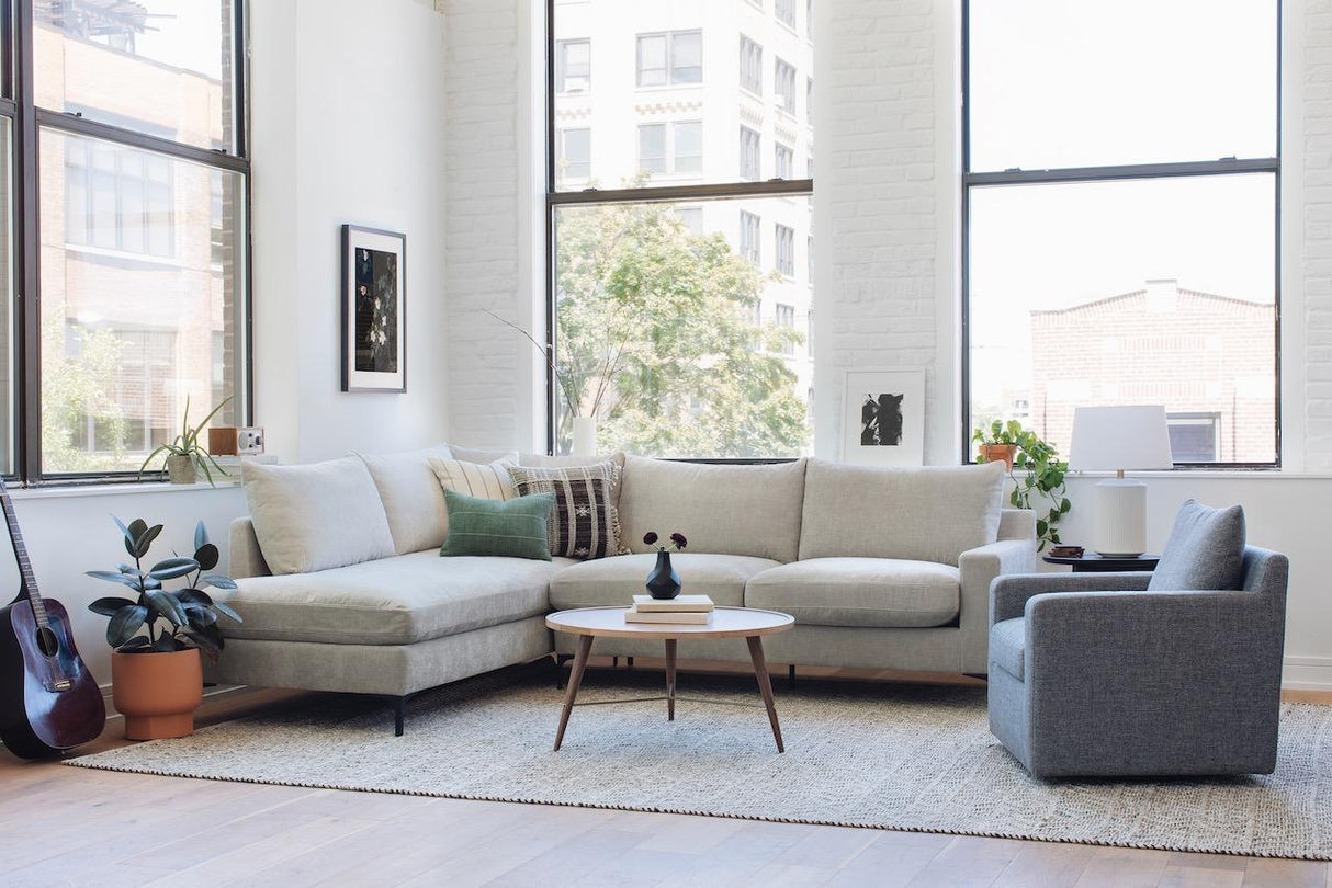 A bright, modern living room featuring a spacious Sloan Custom 3-Seat 95" cream sofa in an L-shape, alongside a gray armchair and a round wooden coffee table. The space is complemented by lush plants and a guitar, with large windows allowing natural light to flood in. The minimalist decor is accented by framed art on the walls.