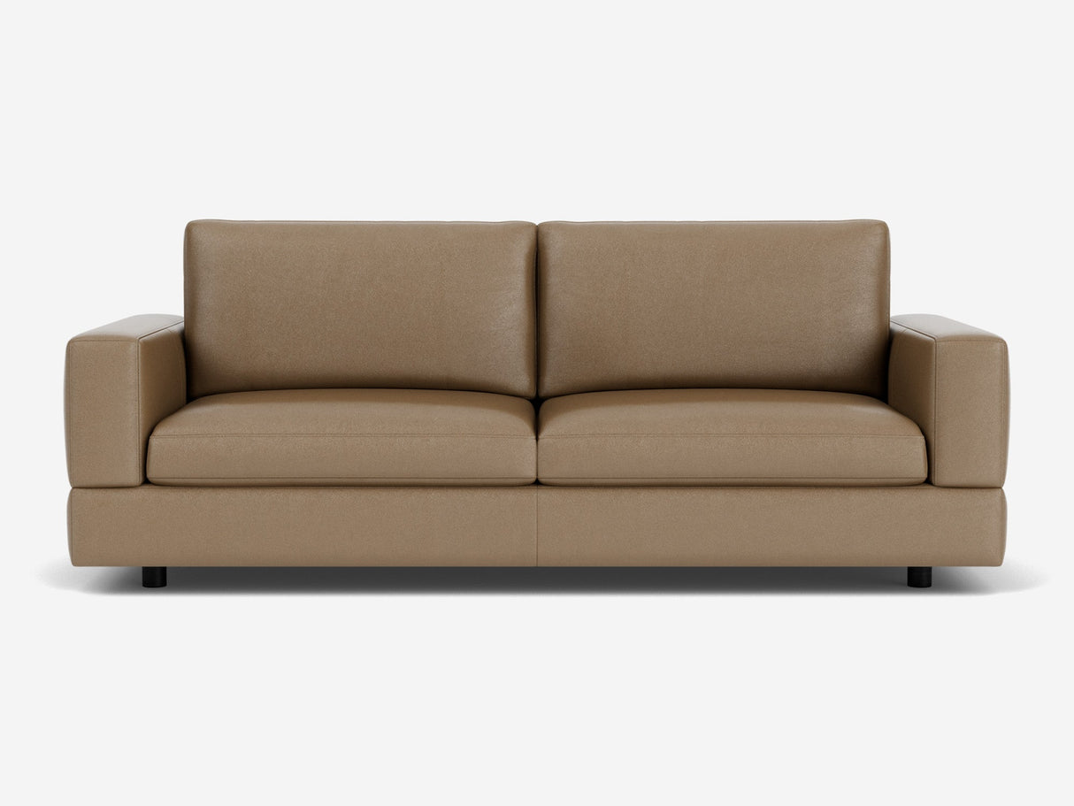 The Stage Track Sofa, featuring a minimalist design with thick cushions and square armrests, is showcased against a white background. Its brown leather upholstery and sleek, contemporary style are enhanced by short black legs.