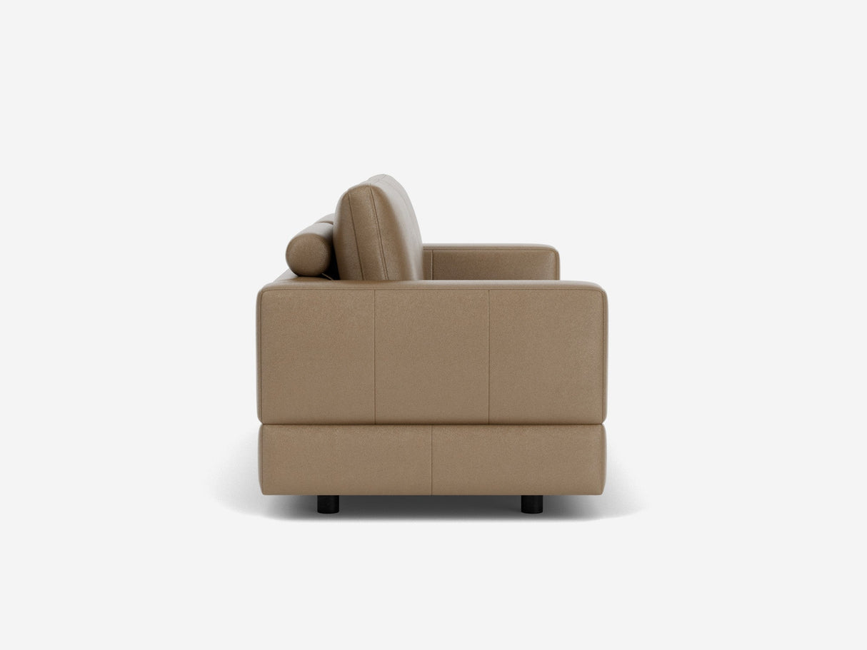 A side view of the Stage Track Sofa, featuring a light brown hue and modern design with clean lines and square armrests. It includes a cylindrical pillow and rests on short, dark legs, all set against a plain white background.