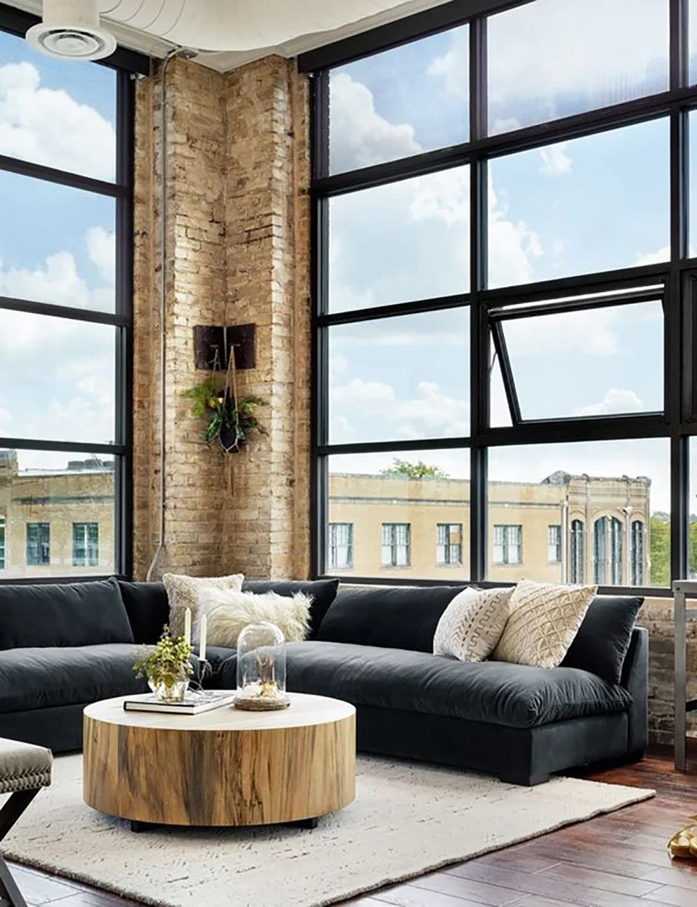 A modern living room with large windows showcasing a cityscape. It features the Decima Sectional Sofa, a dark L-shaped piece adorned with cushions, along with a round wooden coffee table decorated tastefully, and brick walls that allow natural light to fill the space.