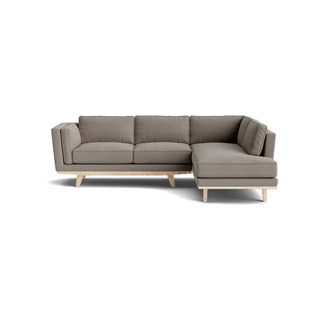 The Kirnik Bumper Sectional in Natural Latex is a contemporary gray sofa with a chaise lounge positioned on the right side, featuring wooden legs and plush cushions, all set against a plain white background.