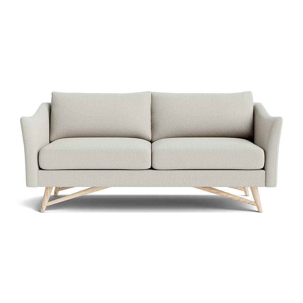 The Gio Sofa in Natural Latex showcases a modern design with its light gray fabric and sleek lines. It features two seat cushions and sits atop a wooden base with angled legs. The sofa's slightly curved armrests contribute to its contemporary and sophisticated look against a white background.