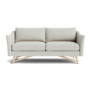 The Gio Sofa in Natural Latex showcases a modern design with its light gray fabric and sleek lines. It features two seat cushions and sits atop a wooden base with angled legs. The sofa's slightly curved armrests contribute to its contemporary and sophisticated look against a white background.