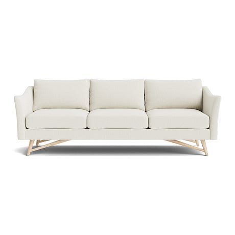 The Gio Sofa in Natural Latex is a modern cream-colored piece, fitted with three cushions and wooden legs. It features angled arms and a minimalist design, all set against a white background.