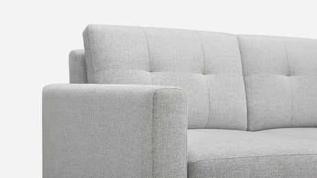 Close-up of the Nomad Fabric Sofa in light gray, showcasing its tufted backrest and sleek, modern lines. The fabric texture is clear, with the image highlighting the armrest and part of the backrest.