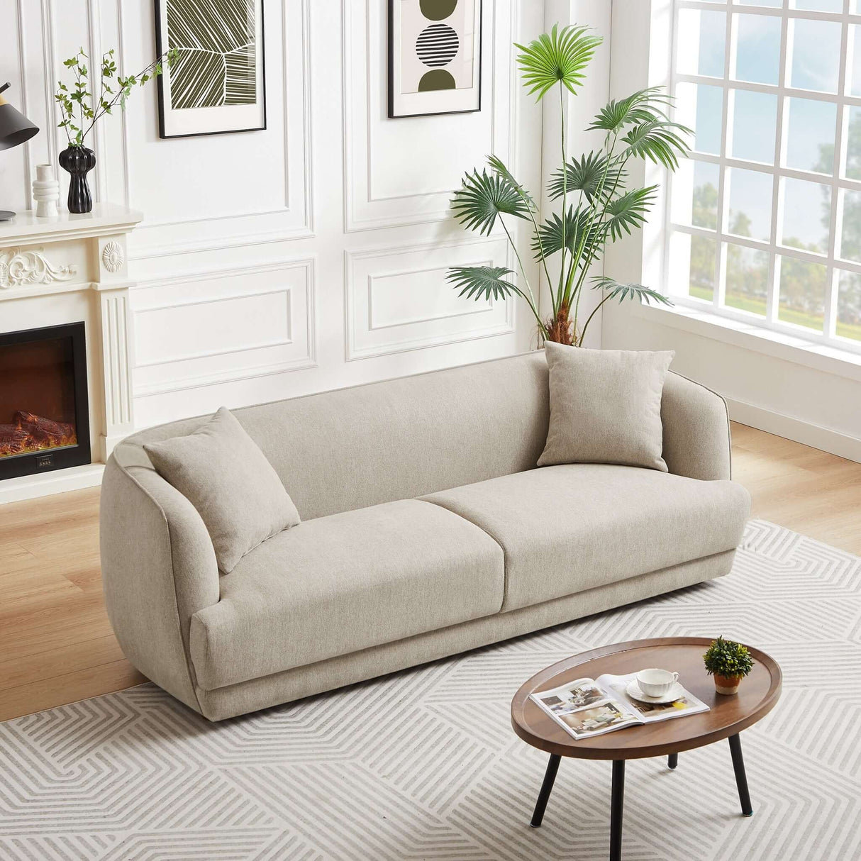 A modern living room features the Larisa Mid-Century Modern Linen Sofa in light beige, complemented by two cushions. There's a round wooden coffee table with magazines and a cup, framed artworks, a tall indoor plant, and a white fireplace with a black vase. A large window completes the decor.