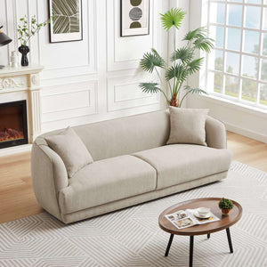 A modern living room features the Larisa Mid-Century Modern Linen Sofa in light beige, complemented by two cushions. There's a round wooden coffee table with magazines and a cup, framed artworks, a tall indoor plant, and a white fireplace with a black vase. A large window completes the decor.