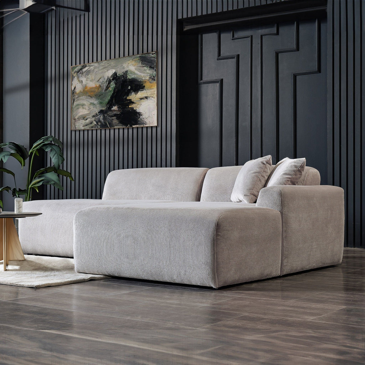 A modern living room showcases the Mar - High-Quality Sectional Sofa in plush gray with two cushions, positioned on a dark wood floor. Nearby is a small round coffee table, while an abstract painting adorns the textured wall and a potted plant adds greenery.