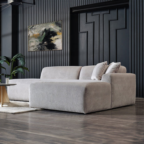 A modern living room showcases the Mar - High-Quality Sectional Sofa in plush gray with two cushions, positioned on a dark wood floor. Nearby is a small round coffee table, while an abstract painting adorns the textured wall and a potted plant adds greenery.
