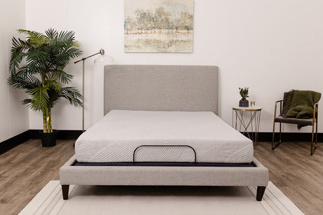 A modern bedroom featuring a light gray upholstered bed with the 8" Comfort Series Firm Gel Memory Foam Tight Top Mattress, wall art above, a potted plant and floor lamp on the left, side table with plant, and a chair draped in a green blanket on the right.