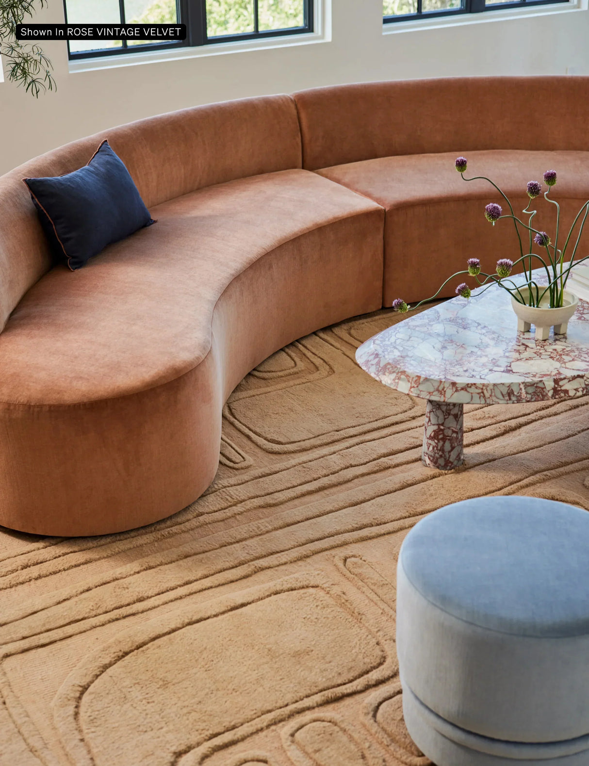 A cozy living room featuring the Cara Sectional Sofa, a curved design upholstered in rose-colored velvet with a blue cushion. A marble coffee table adorned with purple flowers in a vase complements the setup, while a textured beige carpet adds warmth to the space. Large windows in the background bring in natural light.
