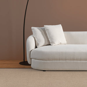 A modern living room features the Chandler - Comfortable Sectional Sofa in white, with two matching cushions against a brown wall. Nearby stands a sleek, curved black floor lamp on light tan carpet and wooden flooring.