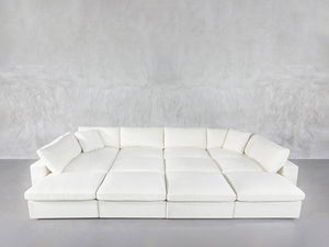 A spacious 12-Seat Deep Modular Daybed with plush cushions and wide seats sits against a light gray textured wall in a minimalist room featuring a concrete floor.