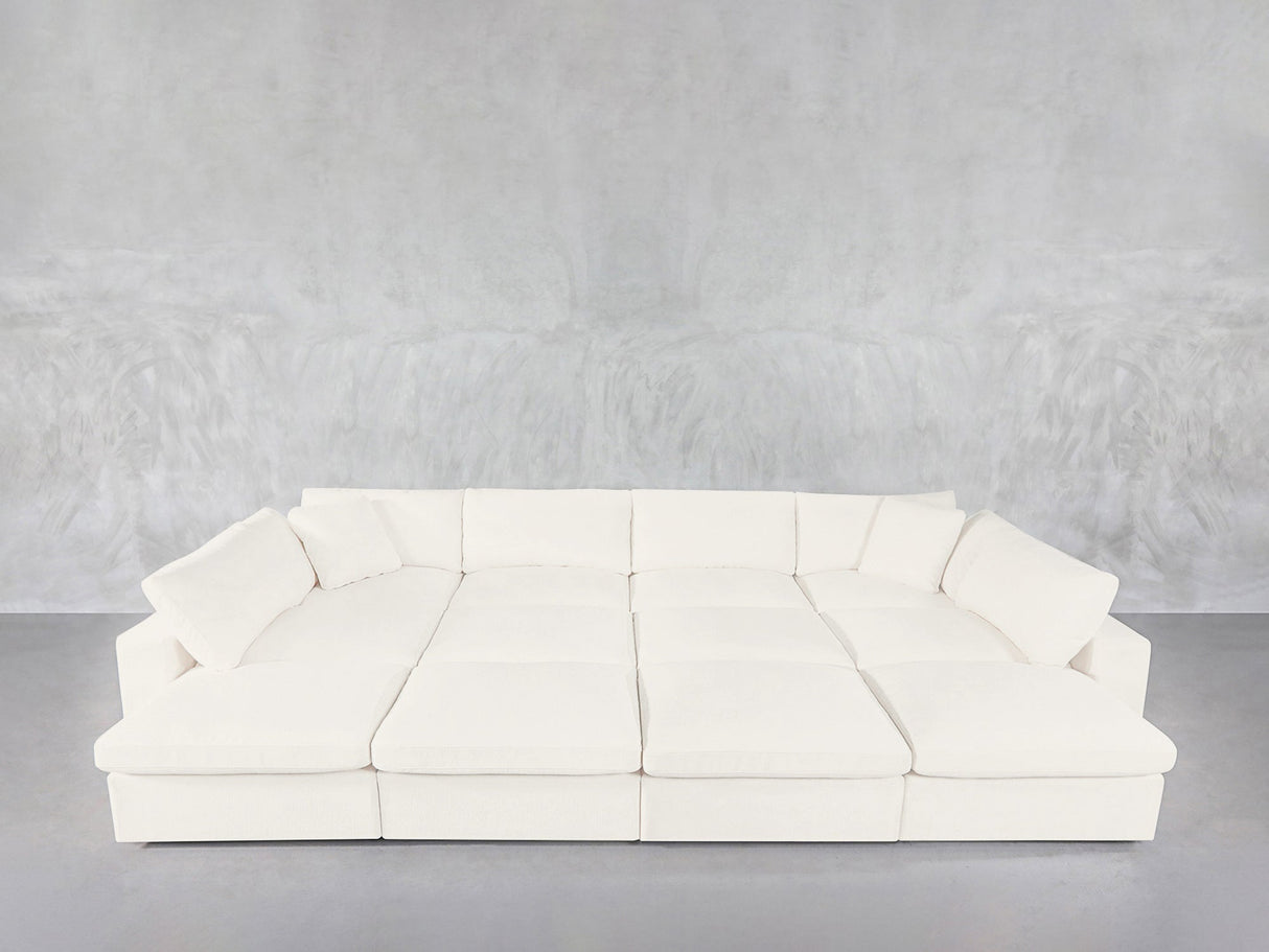 A spacious, modern white 12-Seat Deep Modular Daybed with wide seats and numerous cushions sits against a plain light gray wall and floor.