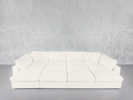 A spacious, modern white 12-Seat Deep Modular Daybed with wide seats and numerous cushions sits against a plain light gray wall and floor.