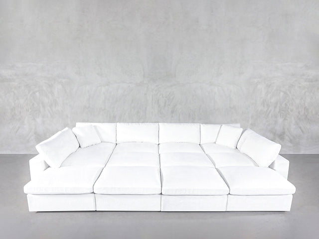 A 12-Seat Deep Modular Daybed with white upholstery and multiple cushions sits against a plain light gray textured wall on a gray floor.
