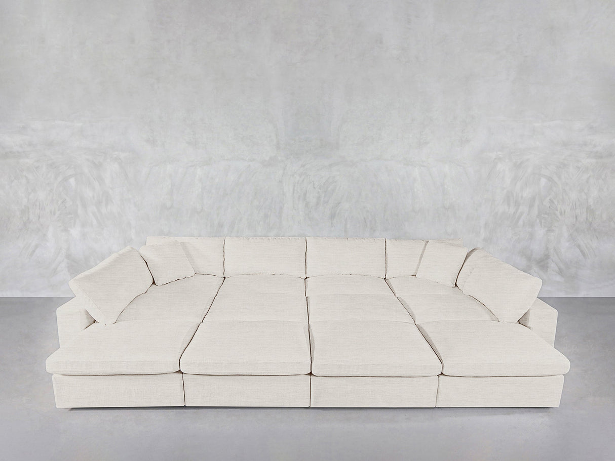 The 12-Seat Deep Modular Daybed with light beige upholstery and matching cushions is set against a textured light gray wall on a smooth gray floor.