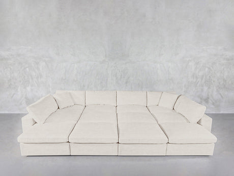 The 12-Seat Deep Modular Daybed with light beige upholstery and matching cushions is set against a textured light gray wall on a smooth gray floor.