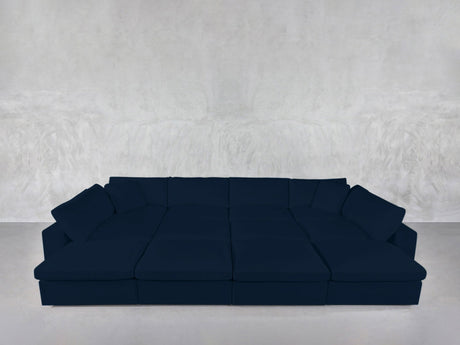 The 12-Seat Deep Modular Daybed, a spacious and modern dark blue sectional with wide seats and multiple cushions, is placed against a simple light gray textured wall and floor.