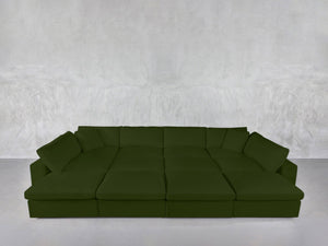 The 12-Seat Deep Modular Daybed, in deep green with multiple seat cushions and back pillows, stands against a light gray textured wall in a minimalistic room with a pale floor.