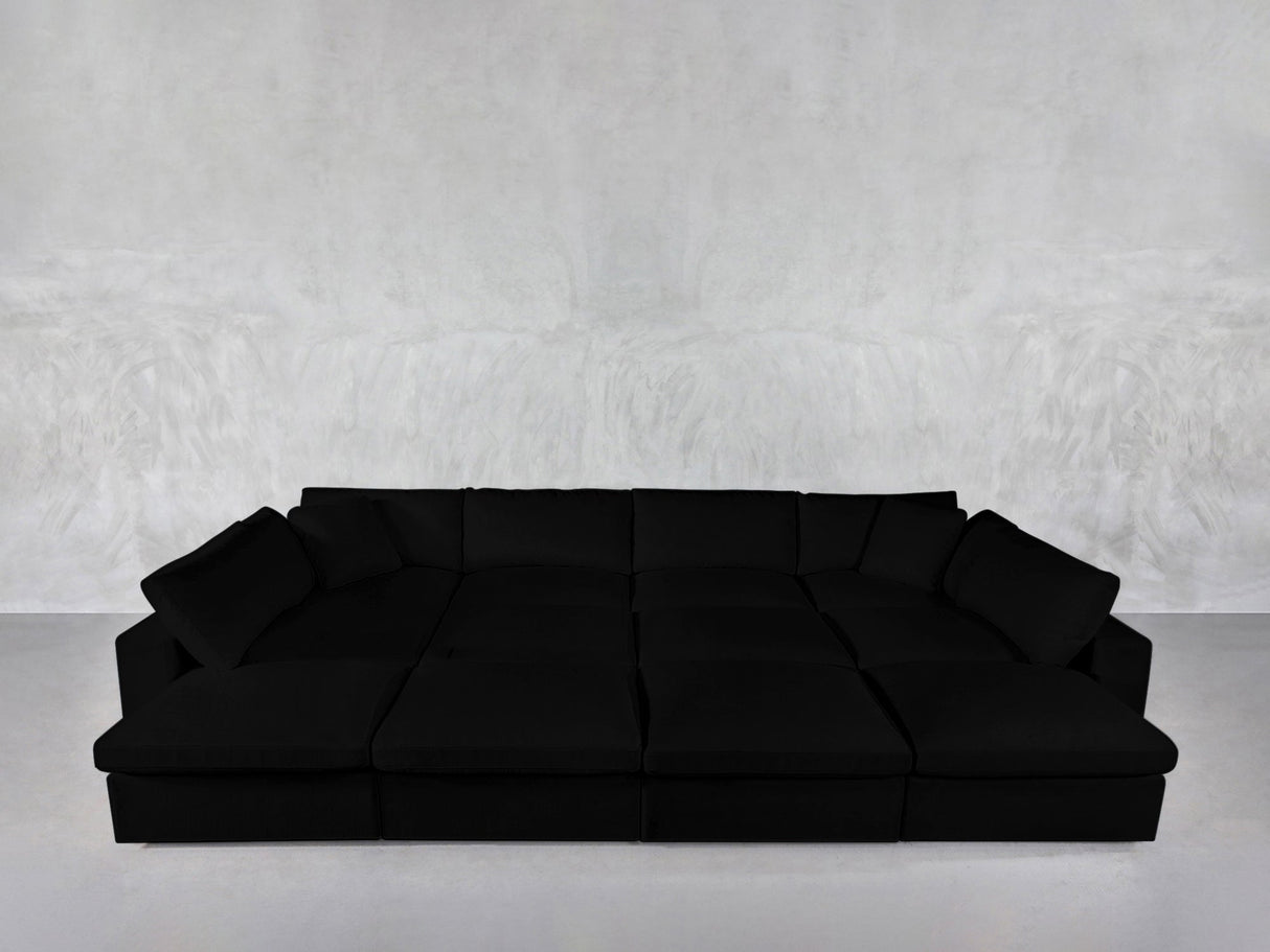 A spacious, modern 12-Seat Deep Modular Daybed with plush cushions sits against a plain light gray textured wall and a smooth, light-colored floor.