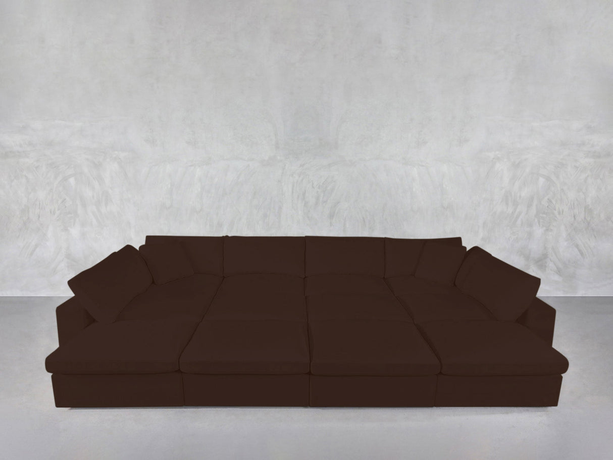 A 12-Seat Deep Modular Daybed in dark brown with multiple cushions is set against a light gray textured wall on a smooth, light-colored floor.