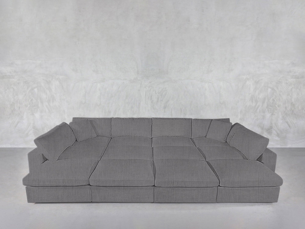 The 12-Seat Deep Modular Daybed, a large modern grey sectional with wide cushioned seats and backrests, is arranged against a plain, light grey textured wall and floor.