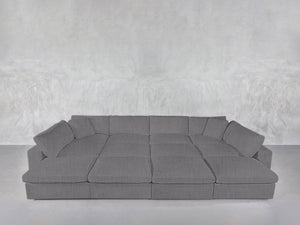The 12-Seat Deep Modular Daybed, a large modern grey sectional with wide cushioned seats and backrests, is arranged against a plain, light grey textured wall and floor.
