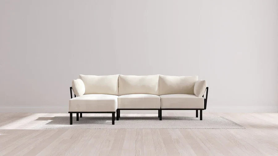 A minimalist, cream-colored sectional sofa with black frame on a light-colored rug in a room with pale wooden floors and a plain white wall. Natural light creates soft shadows on the floor.