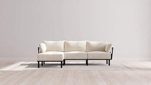 A minimalist, cream-colored sectional sofa with black frame on a light-colored rug in a room with pale wooden floors and a plain white wall. Natural light creates soft shadows on the floor.